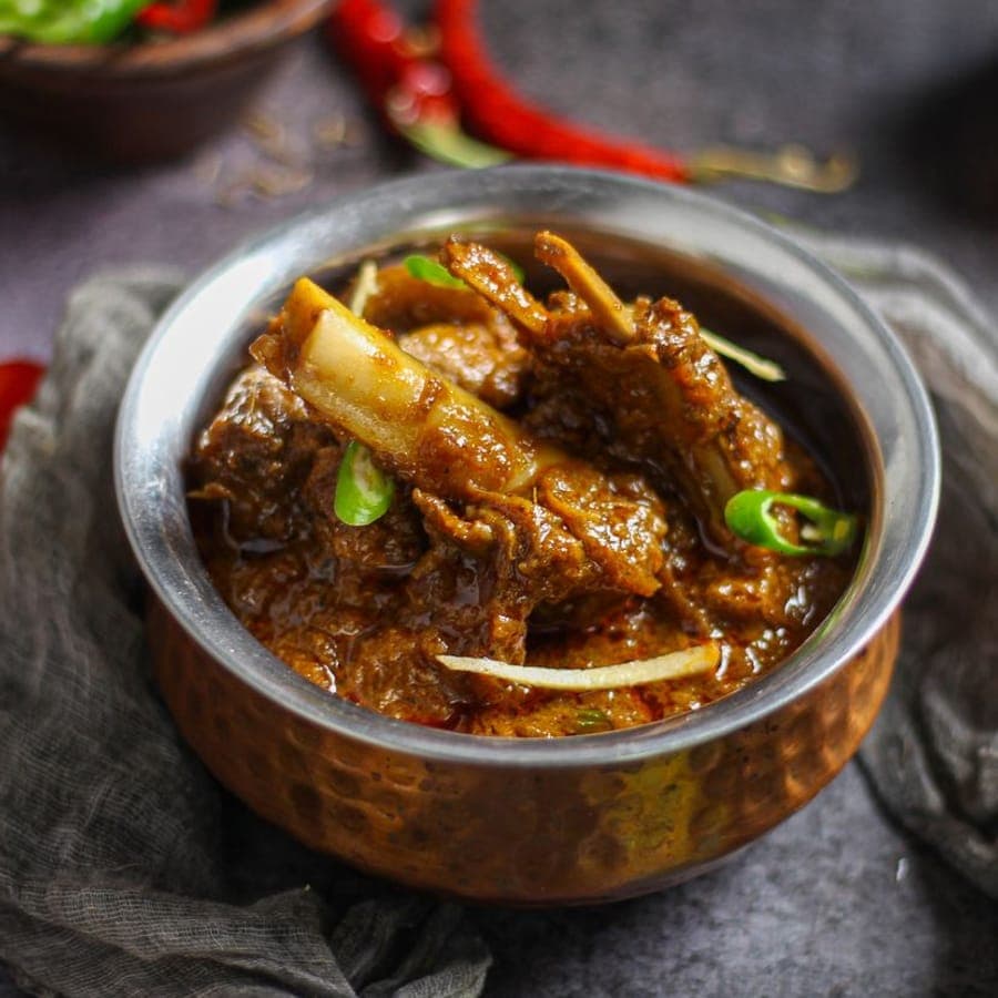 A dark, slow-cooked mutton curry in a hammered copper bowl, topped with slivered ginger and green chilli.
